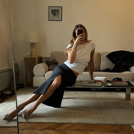 French fashion influencer Anne-Laure Mais taking a mirror selfie in her home in Paris wearing a white t-shirt with an elegant skirt and Gucci heels.