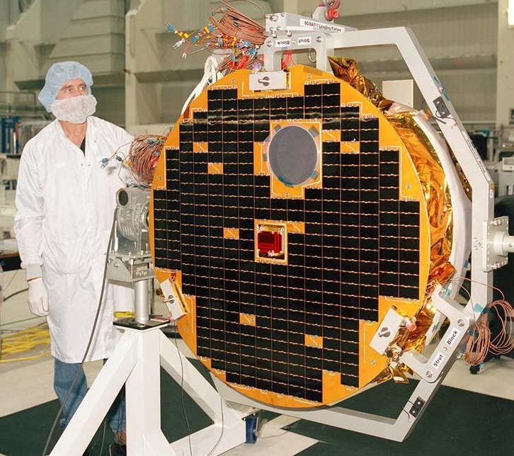 a circular satellite with black solar panels in a laboratory. it is about the same height as a technician standing to the left of it. the technician is wearing a lab coat, hair net and mask to protect the satellite from contamination