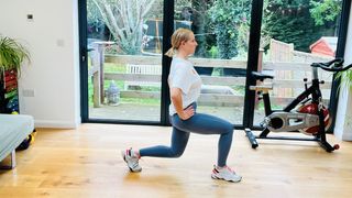 Trainer Maddy Biddulph performs a static lunge hold in her living room. She has stepped forward with her right foot and her right knee is bent. Her left leg remains behind; her left foot is on the floor and the left knee hovers just above the ground. Behind her there is an exercise bike and some large glass sliding doors leading to a garden.