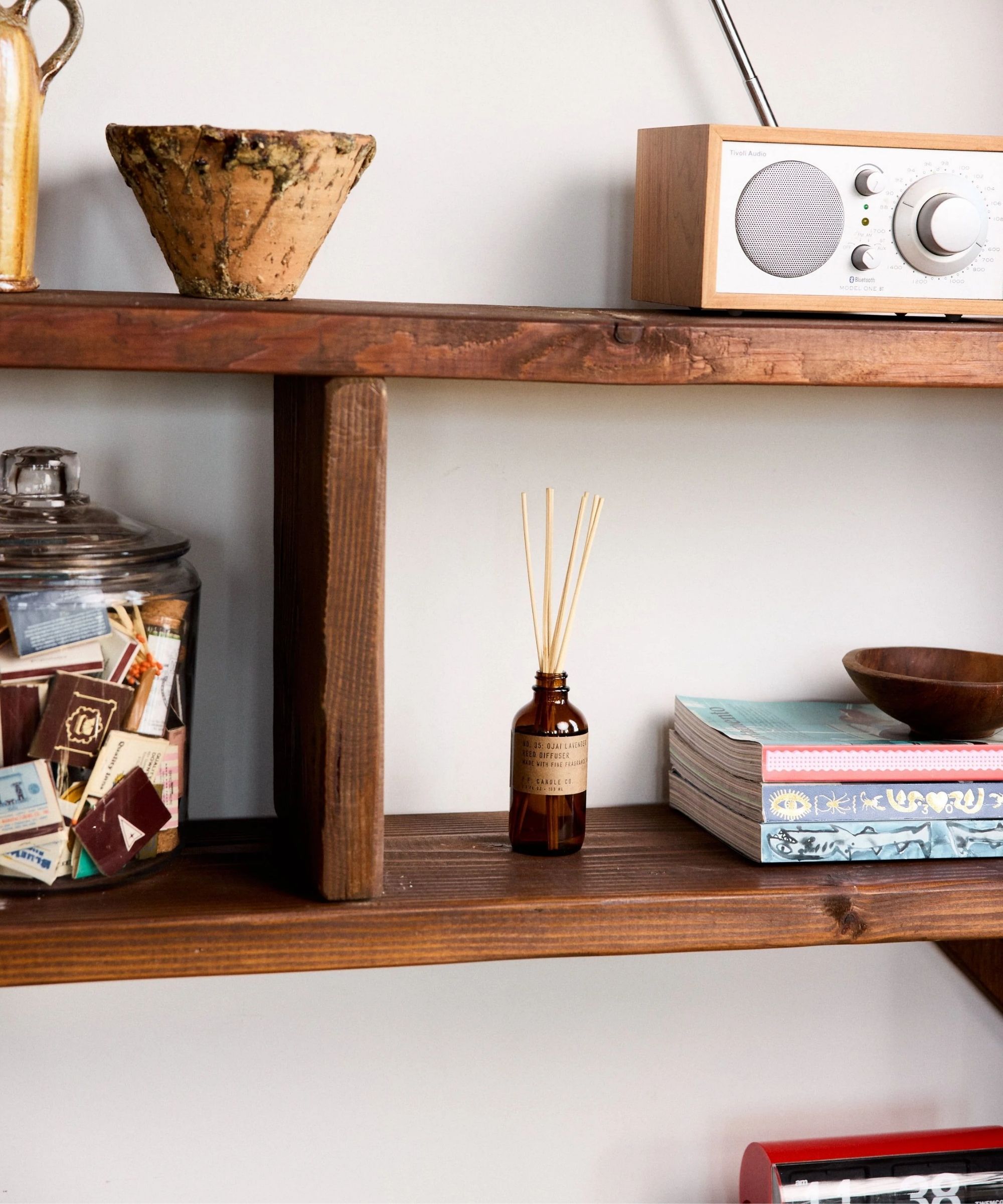 P.F. Candle Co. diffuser on dark wood shelves with books, a wooden bowl, a radio, and a glass jar filled with matchboxes.