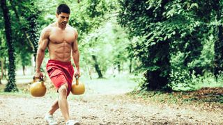 Shirtless muscular person carrying two kettlebells in a forest