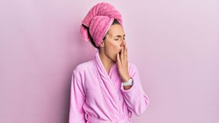 A woman wearing a pink dressing gown with her hair wrapped in a pink towel yawning
