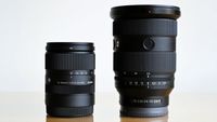 Two black camera lenses, Sigma 18&ndash;50mm and Sony FE 24&ndash;70mm, standing side by side on a reflective surface