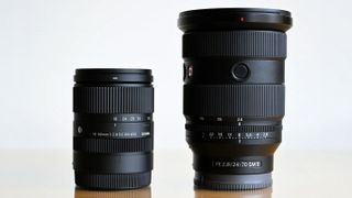 Two black camera lenses, Sigma 18–50mm and Sony FE 24–70mm, standing side by side on a reflective surface
