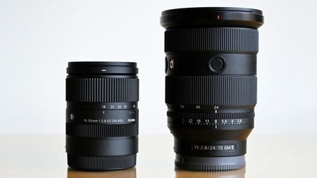 Two black camera lenses, Sigma 18&ndash;50mm and Sony FE 24&ndash;70mm, standing side by side on a reflective surface