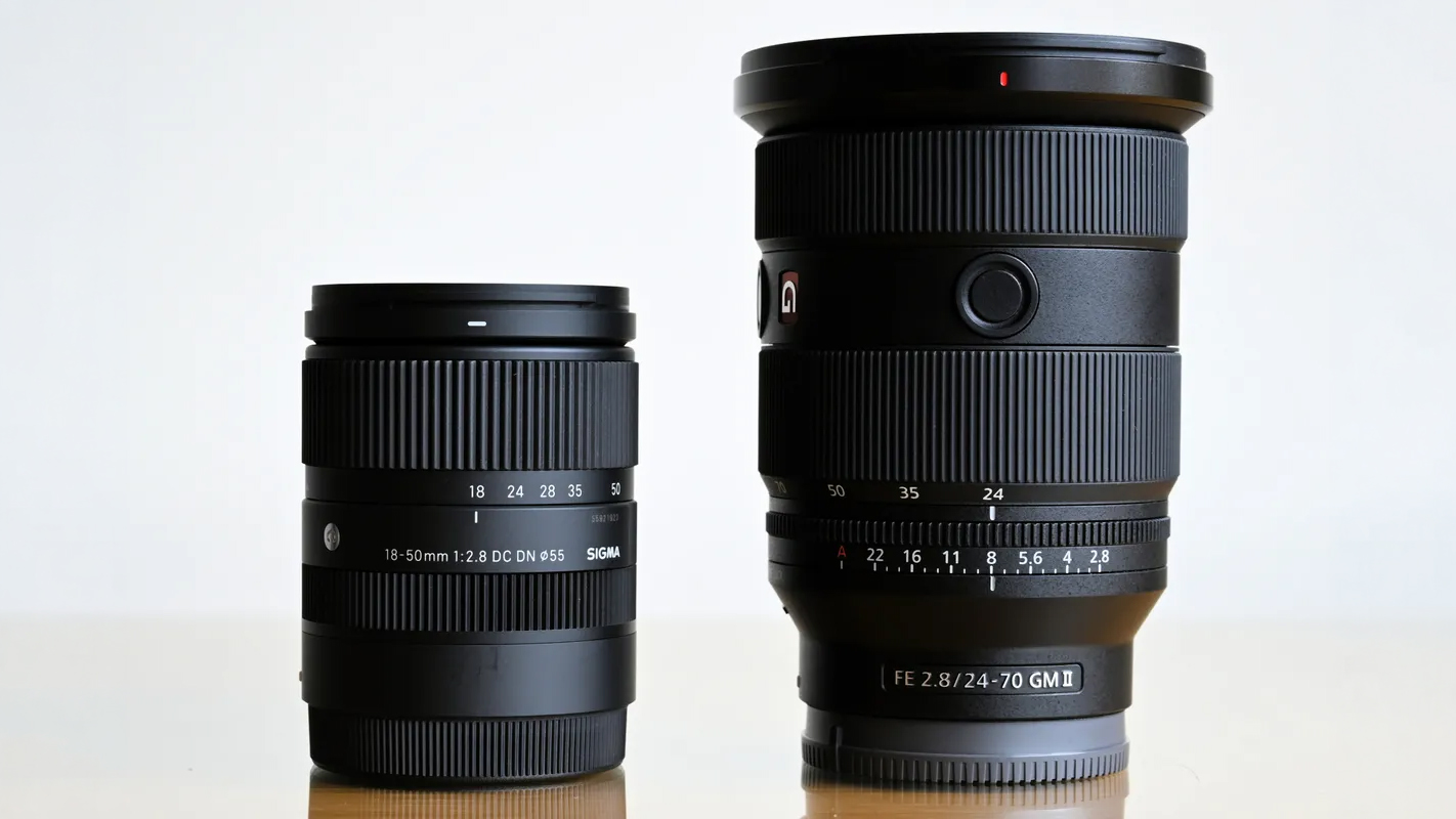 Two black camera lenses, Sigma 18&ndash;50mm and Sony FE 24&ndash;70mm, standing side by side on a reflective surface