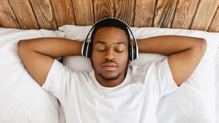 A man lies in bed relaxing, with headphones over his ears, his eyes closed, and his hands behind his head.