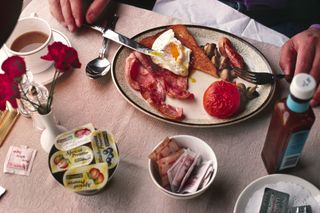 Close-up colour photograph of hands hold- ing a knife and fork and a full English breakfast with a cup of a hot beverage on the table, alongside fruit preservatives, sugar sachets, butter and a bottle of sauce.