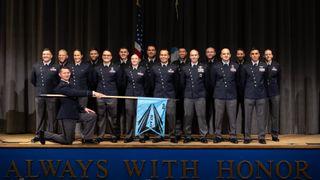 A group of people wearing dark blue shirts and black pants stand on stage with an American flag behind them and an officer kneeling down with a blue flag for the US Space Force placed in front of them.