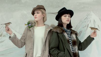 Illustrated snow scene, grey snowy sky and white mountainous background, two female models wearing winter wear, coat and hats, both with an arm out with a small bird perched on their hands
