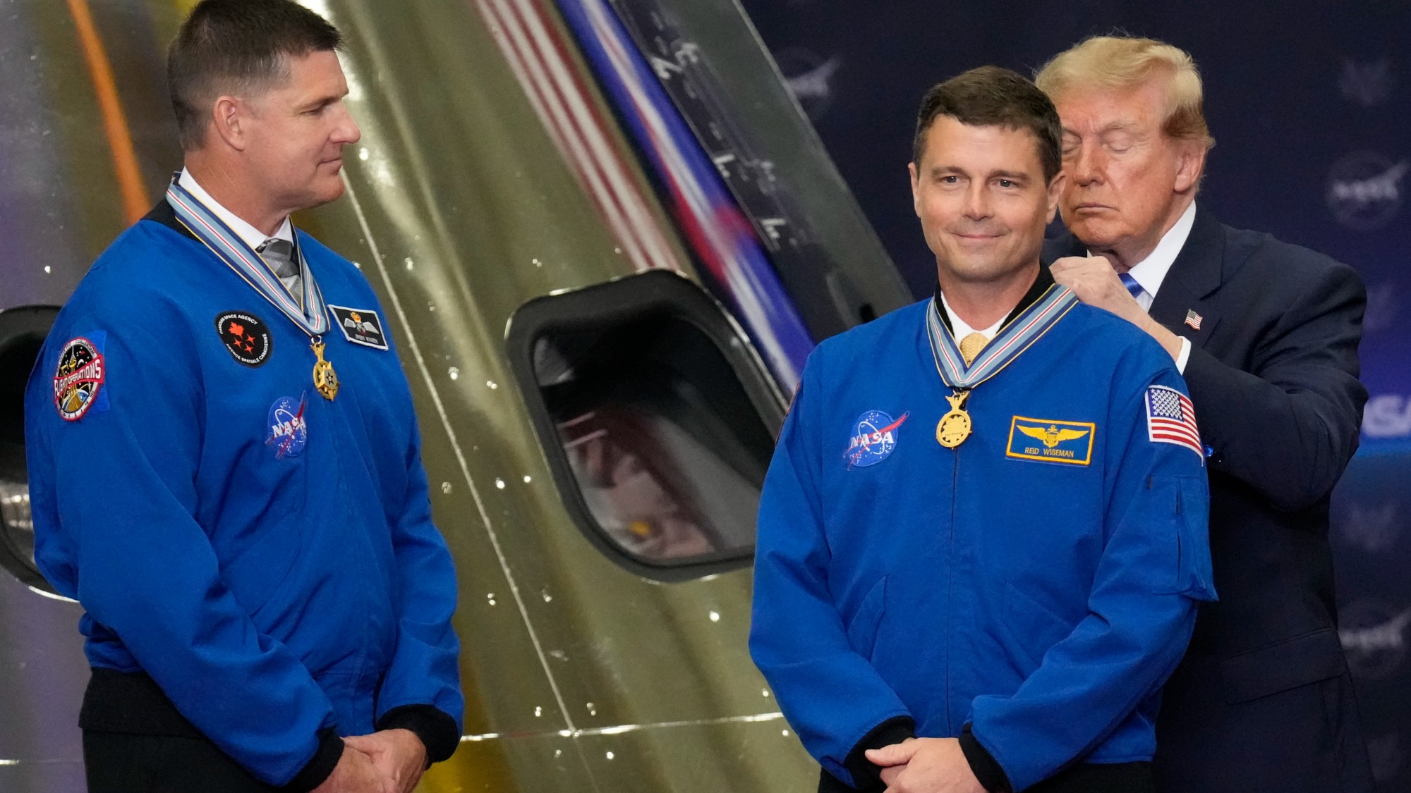 a man in a blue suit gives medals to four people in blue flight suits on a stage with a large sign that reads "NASA"