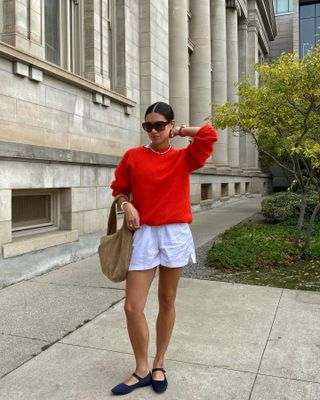 Die Mode-Influencerin Sasha Mei posiert mit einer übergroßen braunen Sonnenbrille, einem leuchtend roten Pullover, einer braunen Wildleder-Hobo-Tasche, weißen Baumwollshorts und schwarzen Mary-Jane-Schuhen aus Samt.