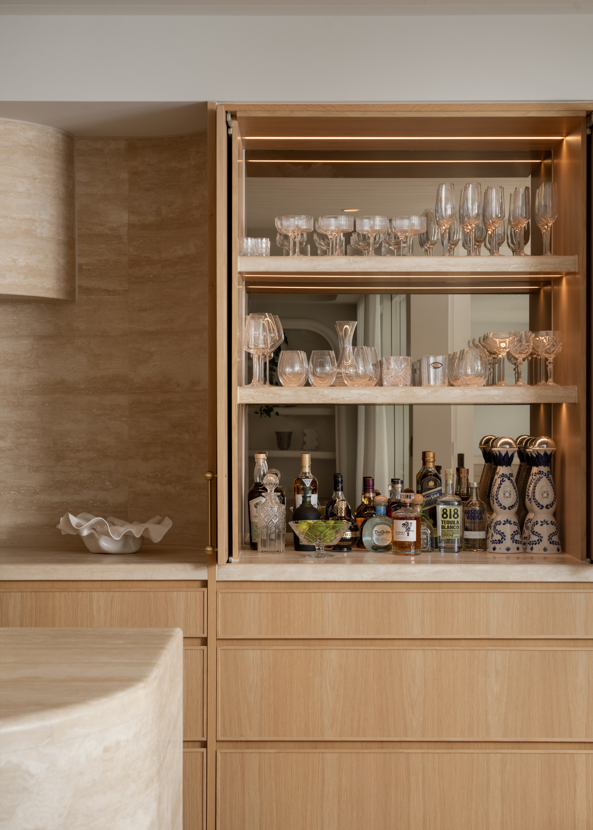 A wood home bar with a mirror by a kitchen with a minimalist marble backsplash