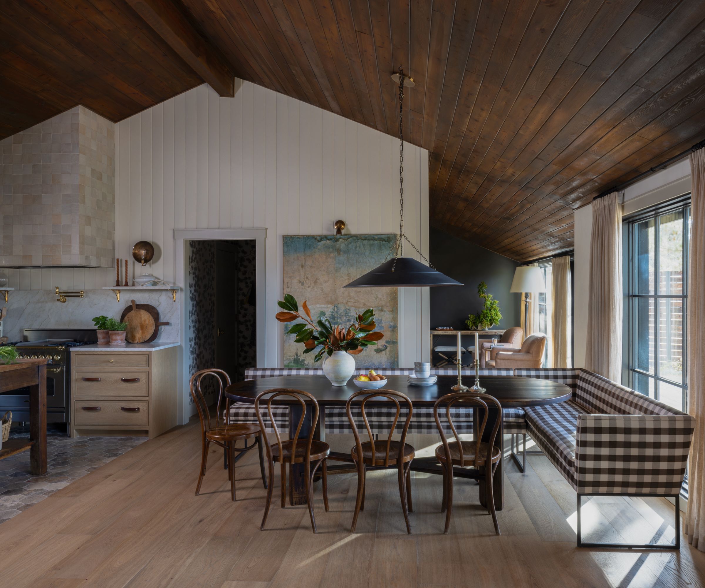 An open-concept kitchen and dining area with a checkered banquette, dark wooden table, and classic chairs. The ceiling is panelled in dark wood and the walls in a light wood.