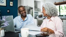 An older couple look happy as they work on paperwork on a laptop at their dining room table.