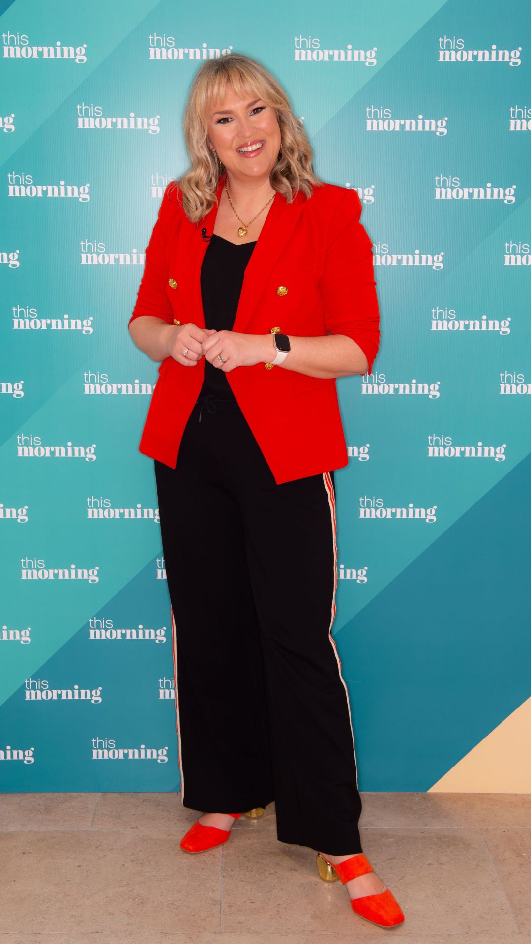 sara davies wearing a red blazer and black side stripe trousers