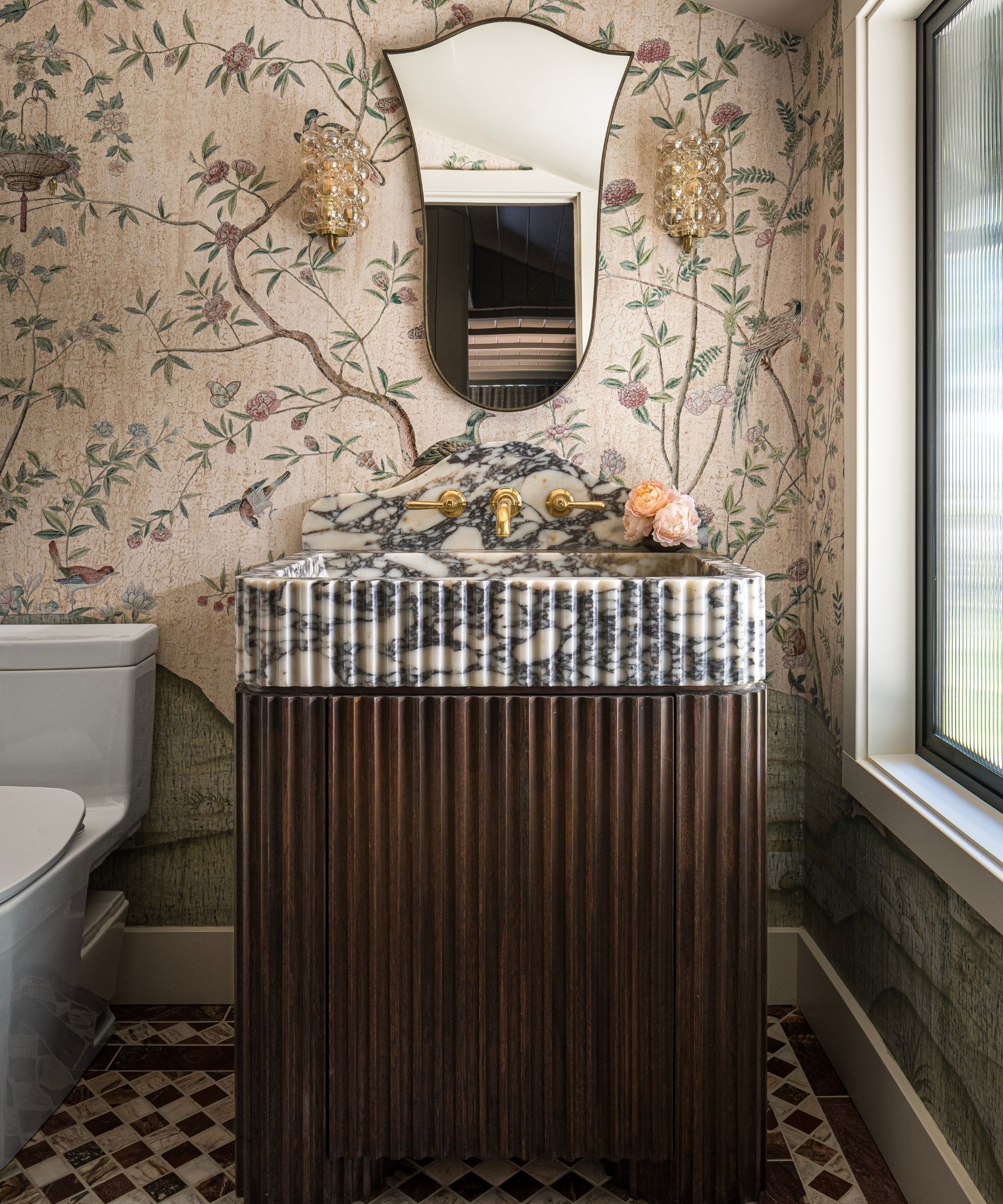 a botanical wallpapered bathroom with marble mosaic floor, fluted wooden vanity with fluted marble sink and vintage mirror