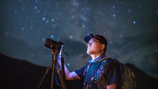 A man with a tripod-mounted camera looking up at the night sky with stars in the background.