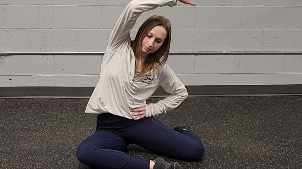 Woman demonstrating stretch. She wears a white long-sleeve top, blue sports leggings and black socks