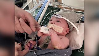 Photo of a baby lying down in a hospital with wires and medical equipment attached it him. He's holding the hand of an adult whose face is out of frame