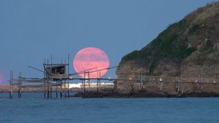 a large pink moon near the horizon with a rocky cliff on the right and a small building on stilts in the water.