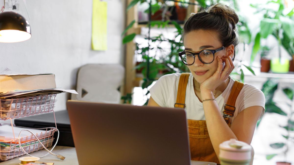 Woman in dungarees sat in a home office, smiling slightly while she looks at her laptop.