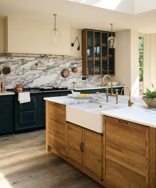 A large wooden island with marble countertops, a white butlers sink, and a gold faucet