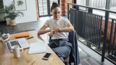 Woman sits at work desk and looks at watch 