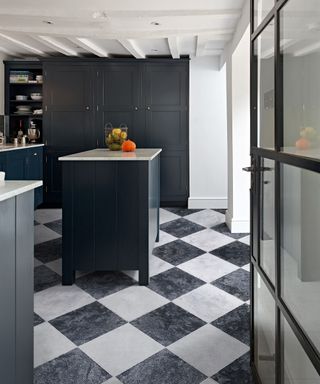 A kitchen with black and white marble checkerboard flooring, a black kitchen island with fruit on top of it, and a black accent wall with a bookshelf to the left