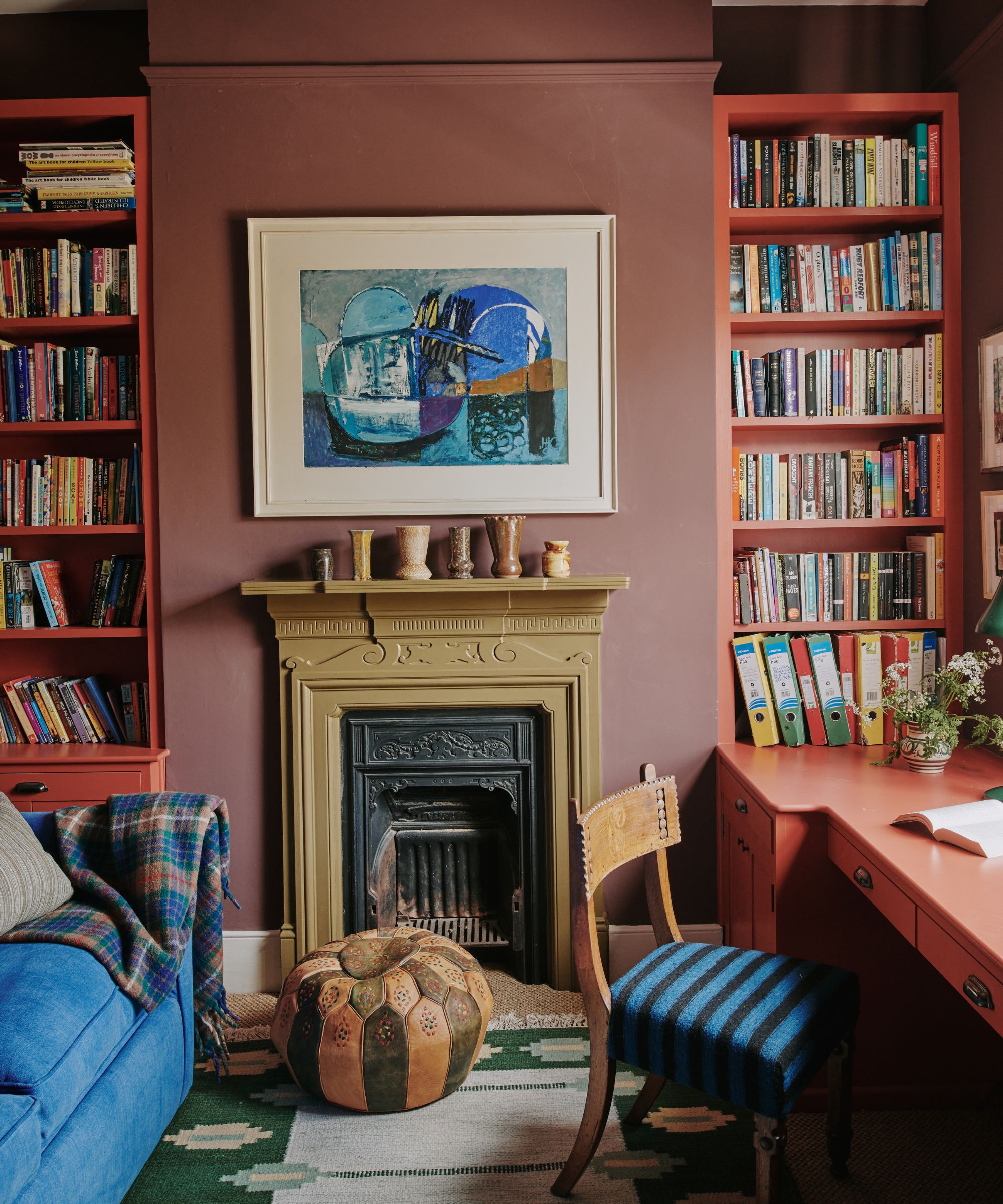 Colourful living room with burgundy walls, red bookcases on either side of the fireplace, a red built-in desk, a vintage upholstered chair, and a blue sofa, with a green woven rug beneath.