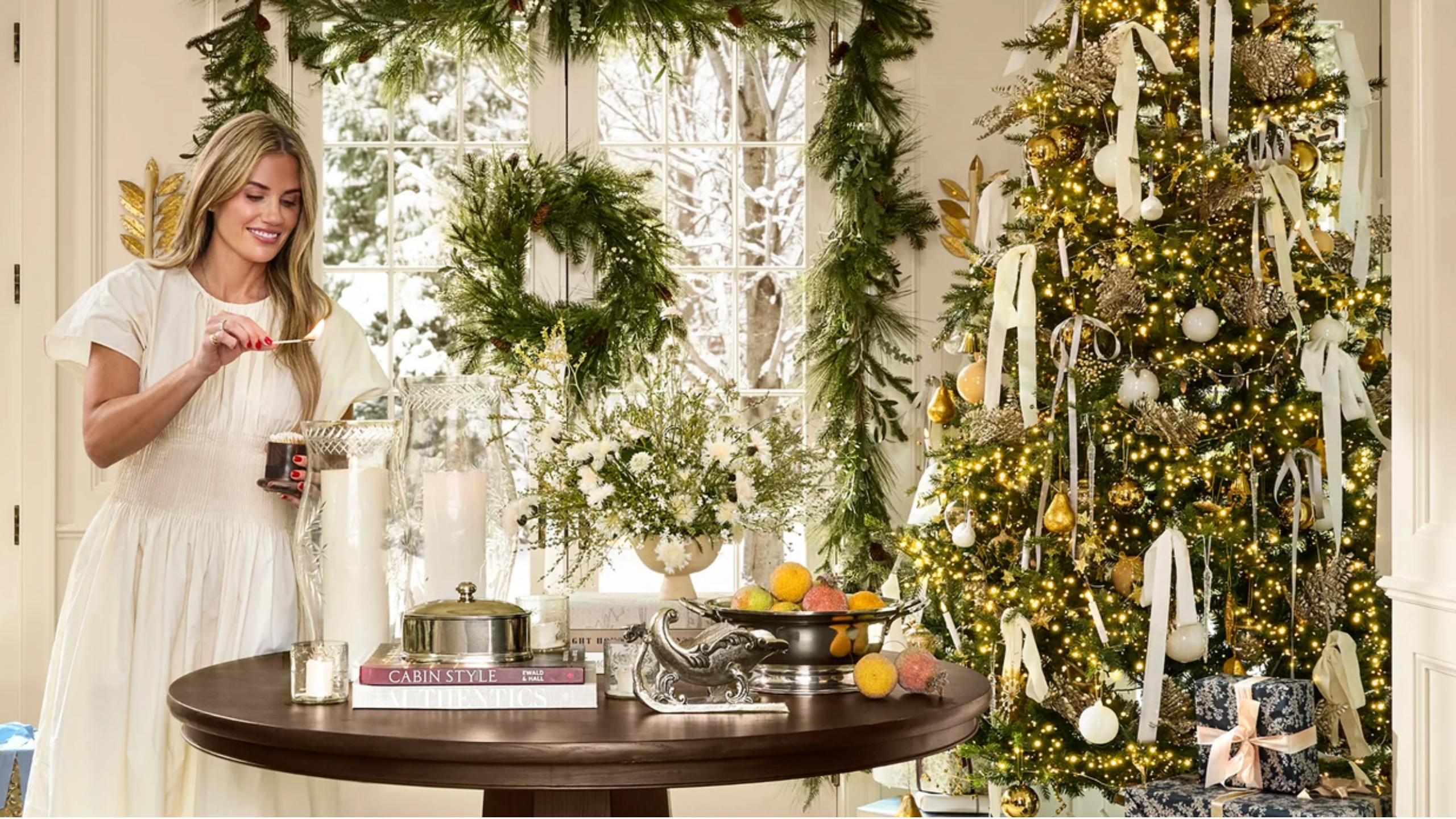 Shea McGee lighting a large pillar candle inside of a festive Christmas living room alongside an ornately decorated tree, wreath, garland, and metallic tabletop accents.