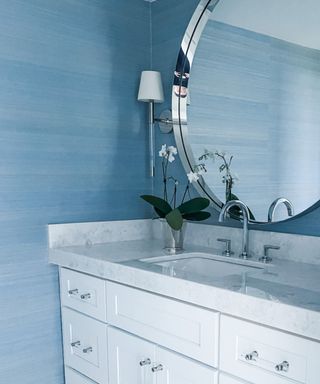 A bathroom with blue walls, a silver circular mirror, a white vanity with gray marble, and blue and white honeycombed flooring
