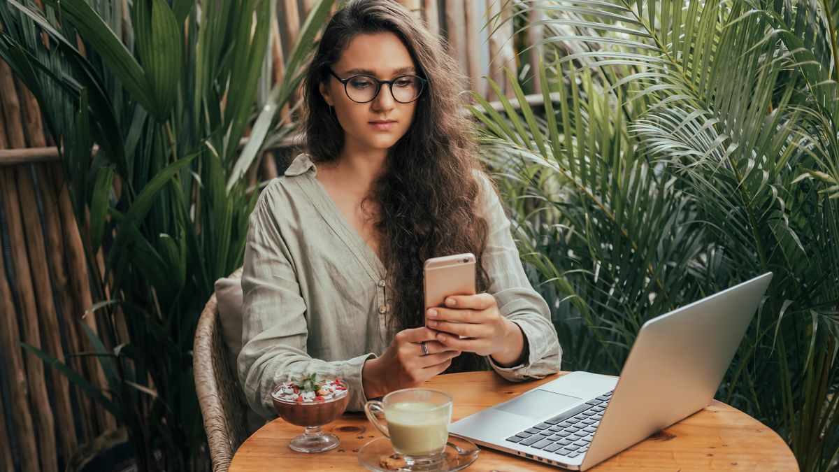 A woman sitting at a table and holding an iPhone, and she has a Mac laptop opened on the table