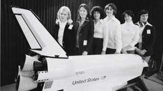 Six black and white women (all white) stand behind a giant model of the space shuttle.