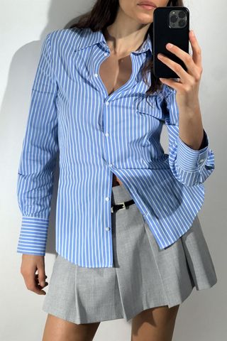 Model wears Fitted Poplin Shirt in blue and white stripes and gray pleated skirt