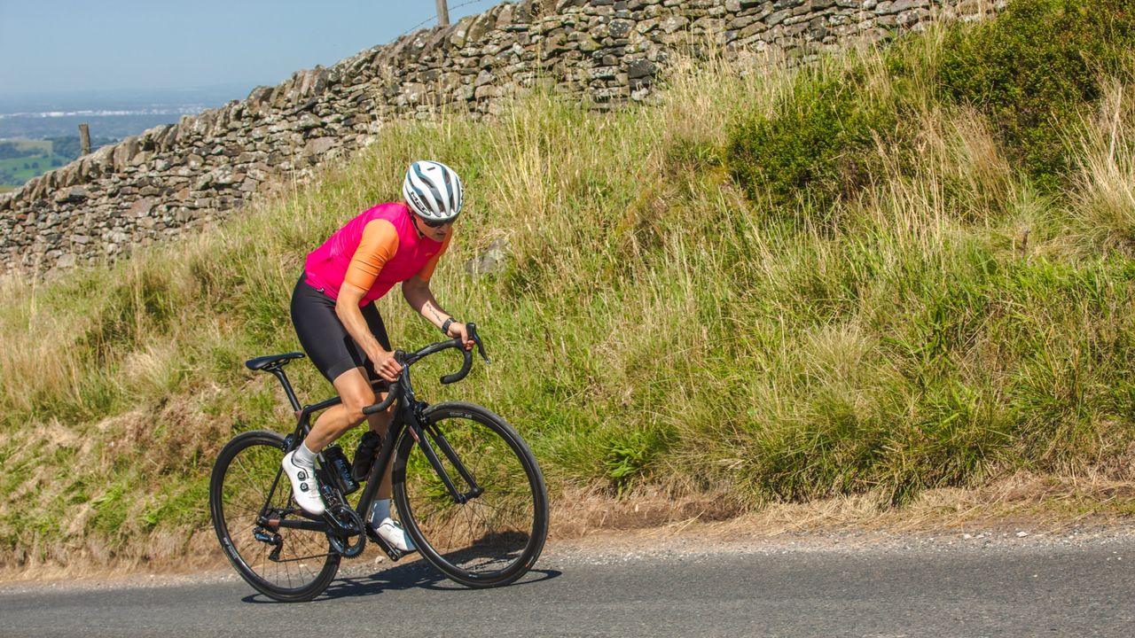 A female cyclist climbs out of the saddle in a pink top and white helmet with greenary in the background