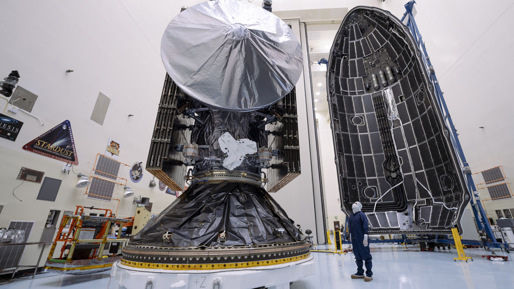 The large Europa Clipper spacecraft has a circular antenna, a man looking at it, and a huge rocket nose cone in the background.