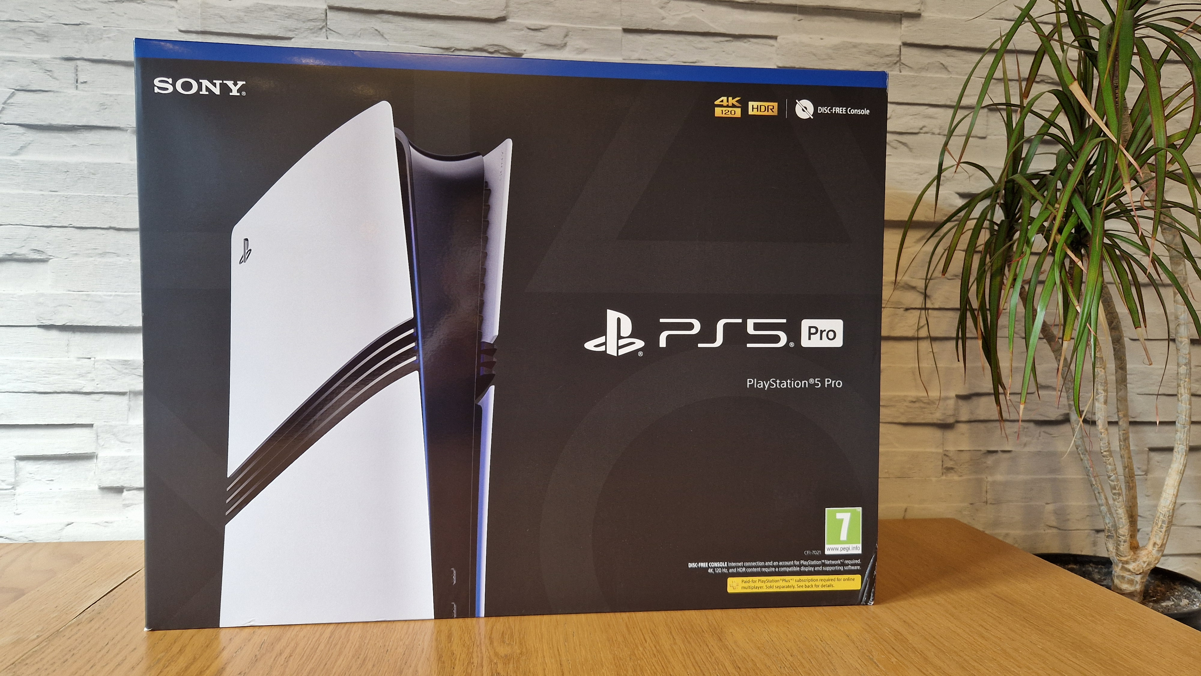 The PS5 Pro box on a wooden desk in front of a white brick background and next to a potted plant