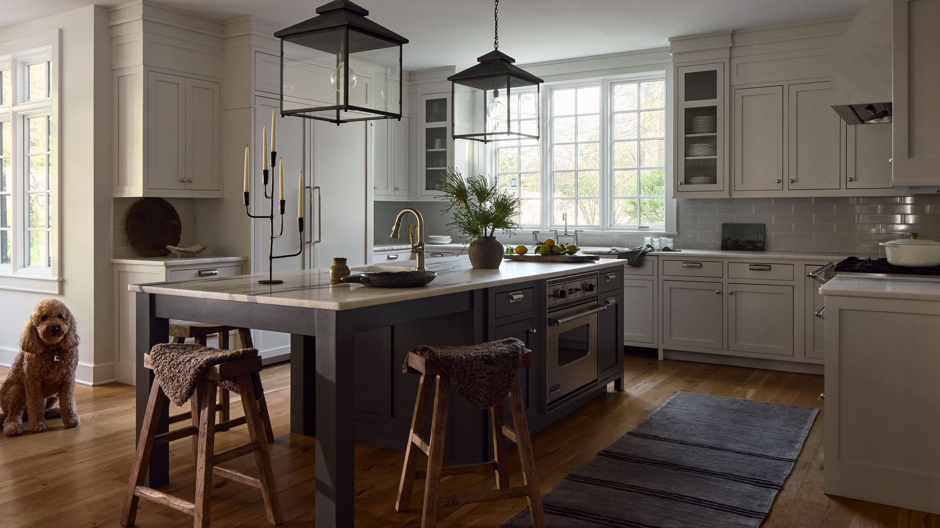 Large kitchen with pale gray wall cabinets and a dark gray island in the center. Two lantern pendant lights hang above and rustic wooden tools provide seating at one end