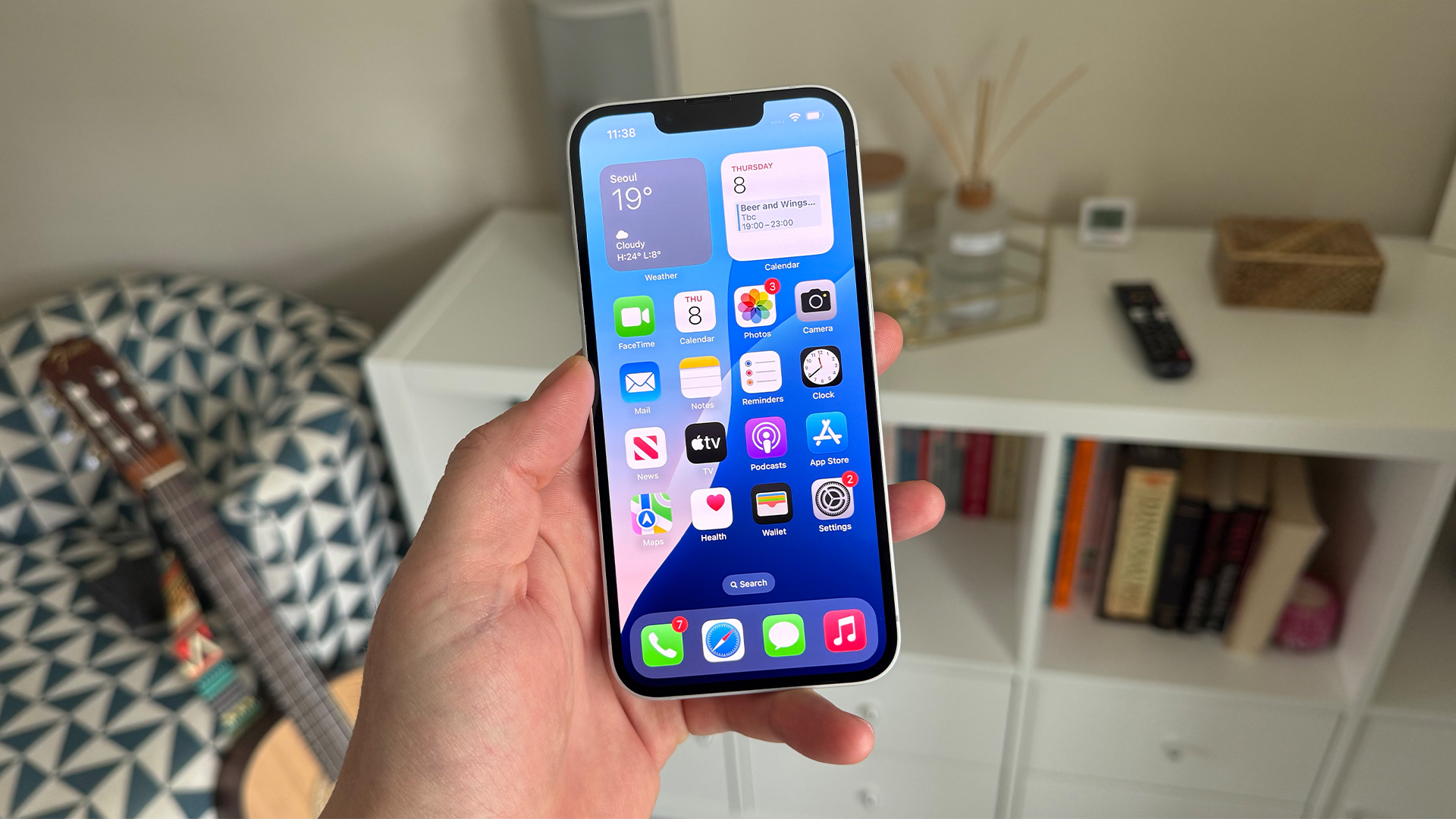 Apple iPhone 16e smartphone held in hand showing apps on screen in front of bookcase and guitar