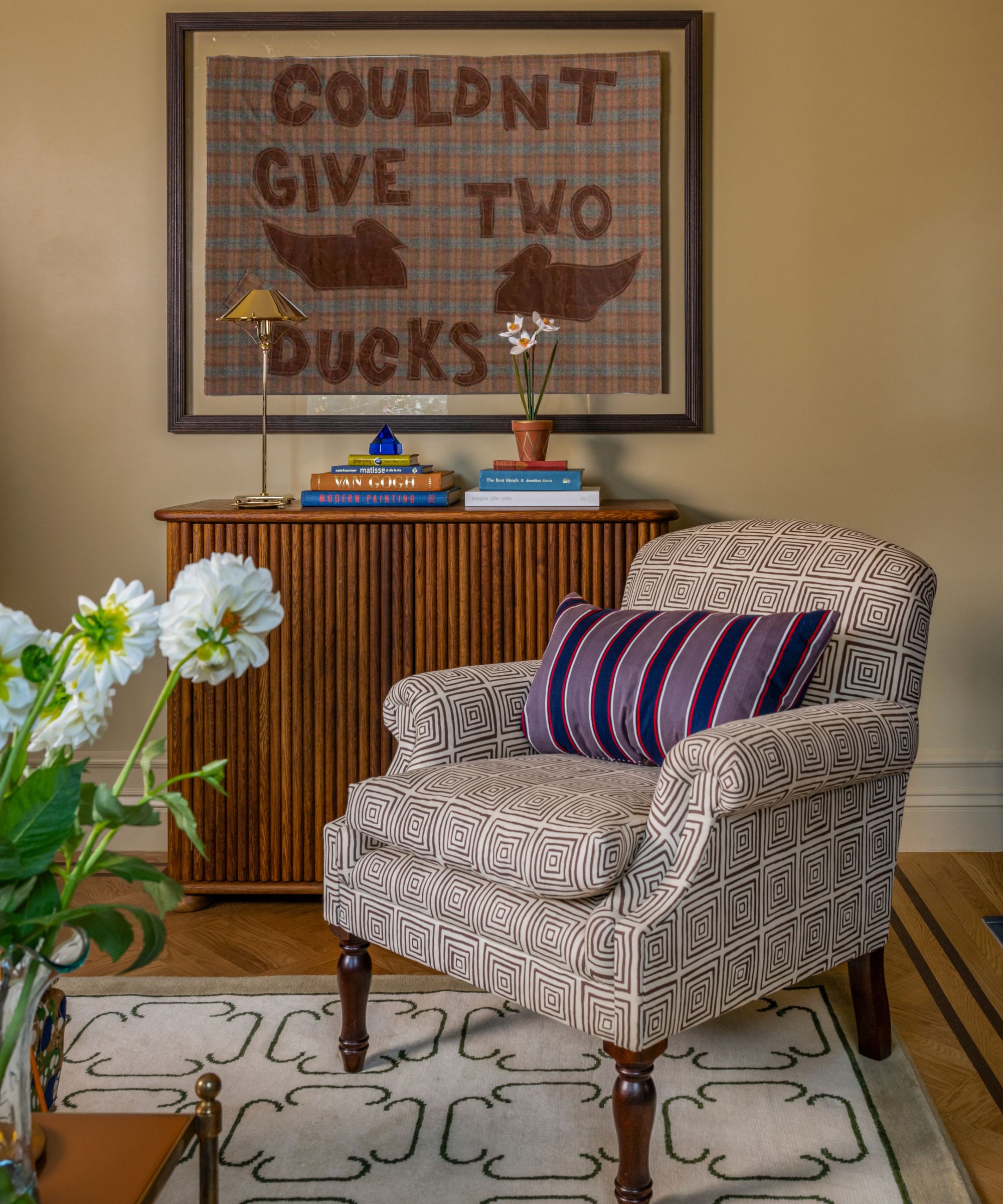 a warm yellow home library corner with a wooden cabinet, geometric printed armchair and cheeky artwork on the wall