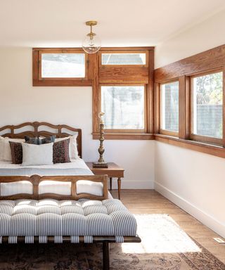 Unusual, wood framed windows across the corner of a cream bedroom with wooden bed and pale white and blue linen.