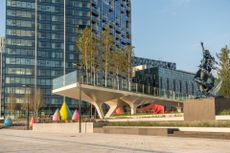 View of The Tide riverside park during the day. The park features a raised walkway, grass, trees, a statue on a plinth and colourful waterdrop-shaped structures. There are glass front buildings nearby