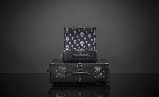 A black suitcase with studs on the front of it with a smaller suitcase on top of it and a bag with skulls printed on it behind them.