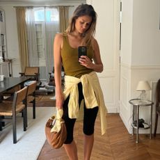 Influencer @annelauremais takes a mirror selfie wearing a khaki tank top, black capris and heeled sandals. She wears a yellow shirt looped around her waist and holds a Chloe bracelet bag.