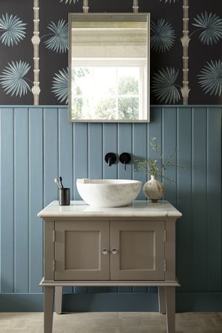 Light filled blue bathroom with panelled blue wall on the bottom and wallpaper on top