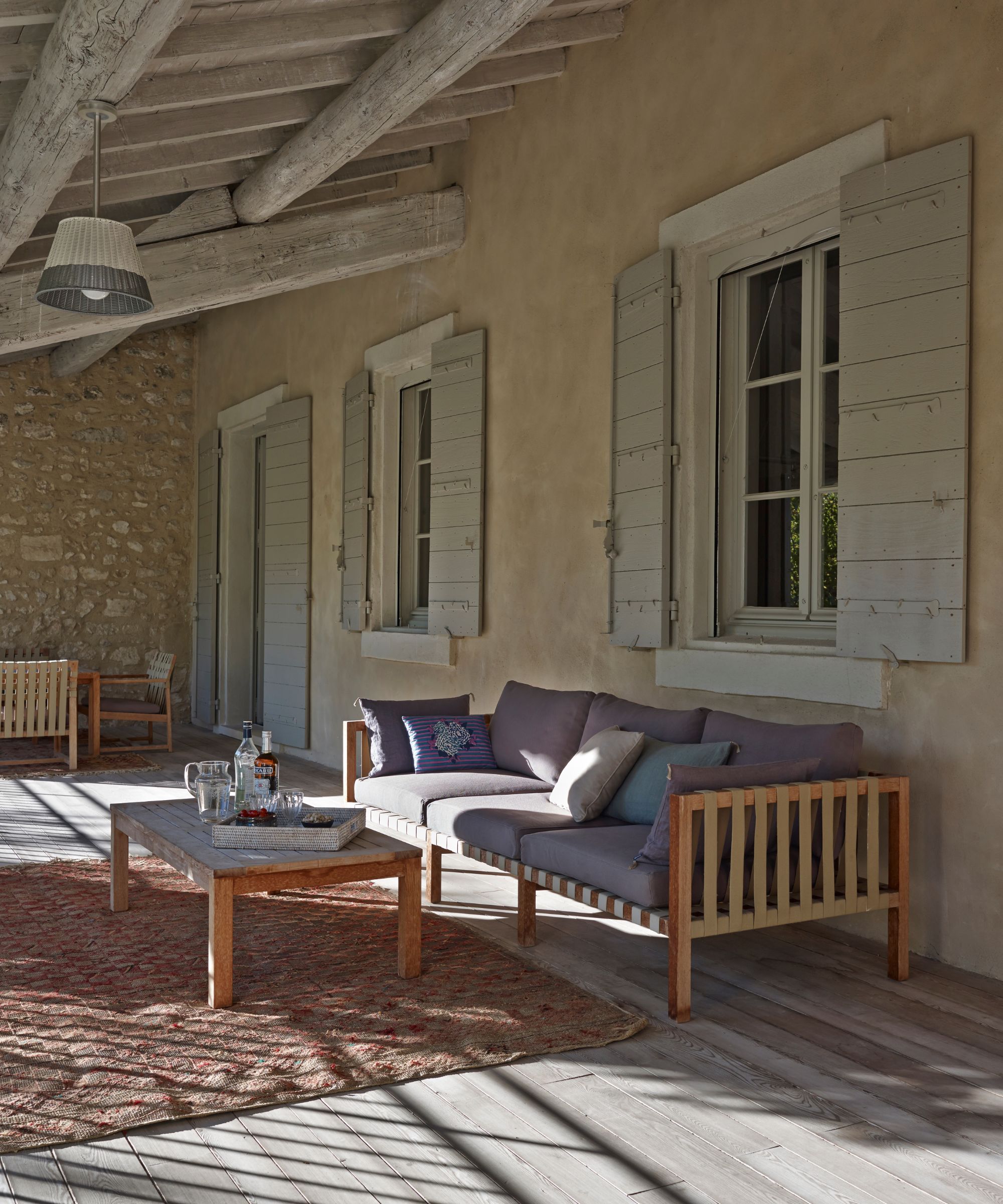 A large porch seating area with shuttered windows, a large wooden sofa with purple cushions, wooden coffee table and large outdoor rug. Above are wooden beams and a light fixture with a white and grey lampshade.
