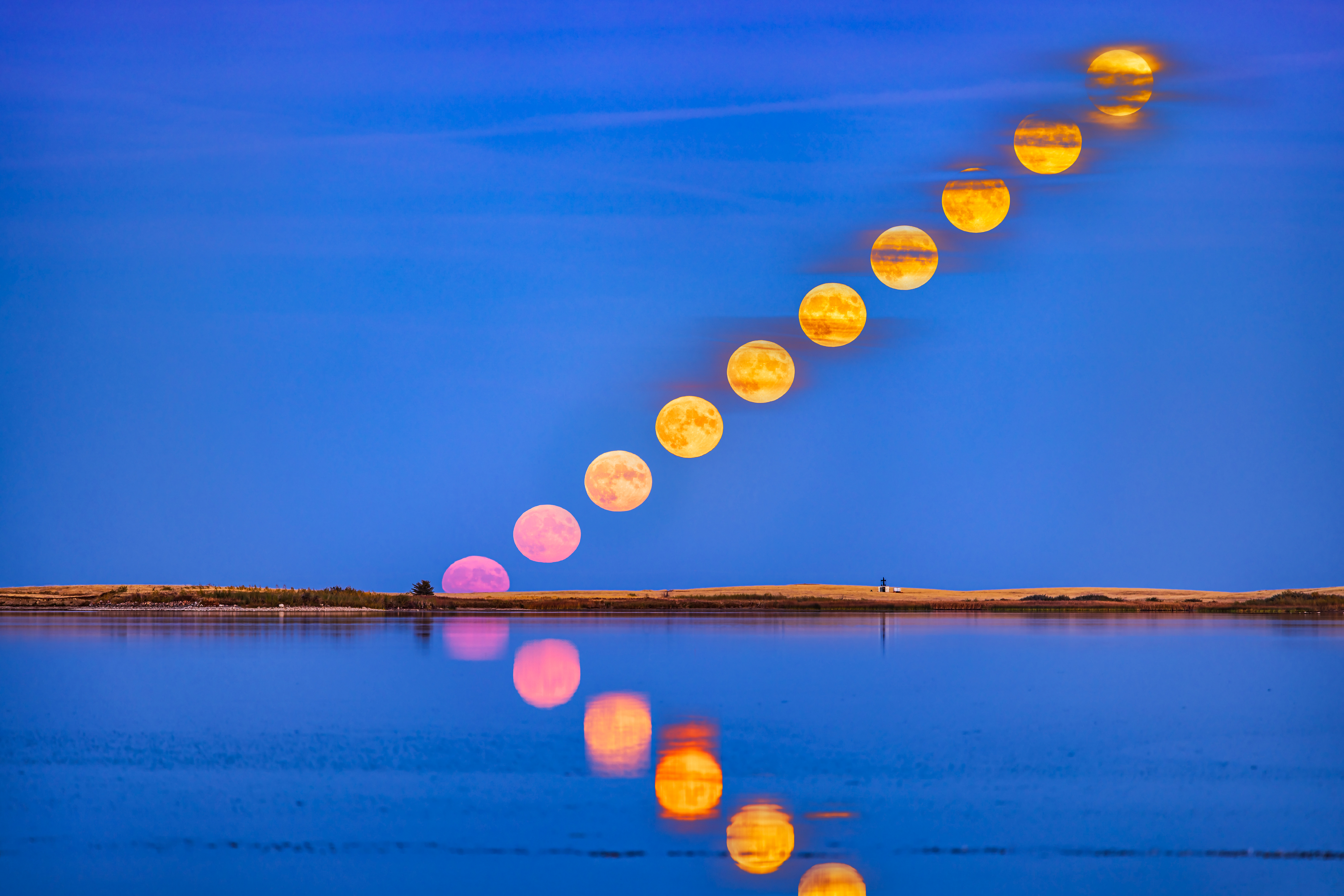 The rising of the full Hunter's Moon of October 9, 2022, with the moon reflected in the calm waters this night at Crawling Lake Reservoir in southern Alberta, Canada. The moon appeared very pink as it rose into the clear sky near the horizo, then turned more yellow as it rose into scattered bands of cloud. (Image credit: Alan Dyer/Stocktrek Images via Getty Images)