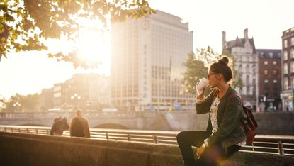 Woman drinking coffee while sitting retailing wall of bridge in city