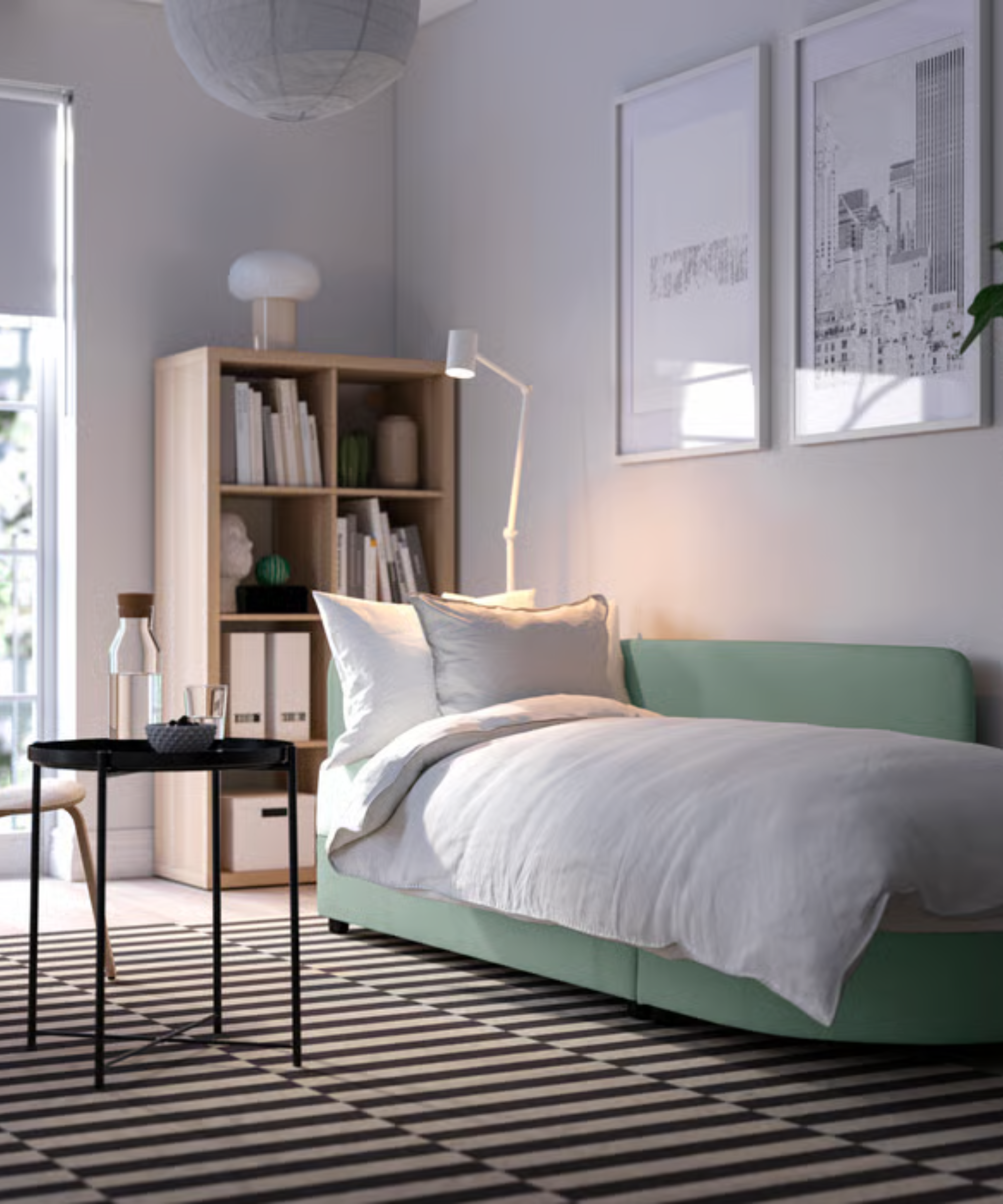 Contemporary gray-green IKEA sleeper sofa styled with white bedding in a neutral living room with a wooden bookshelf, geometrically striped rug, and metal coffee table